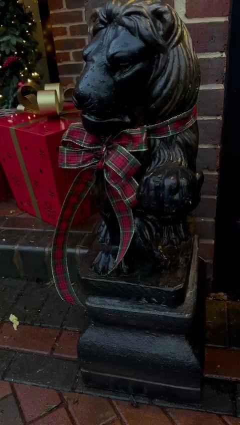A stone lion at an entrance wearing a wide tartan ribbon bow, with wrapped red gift boxes behind it.