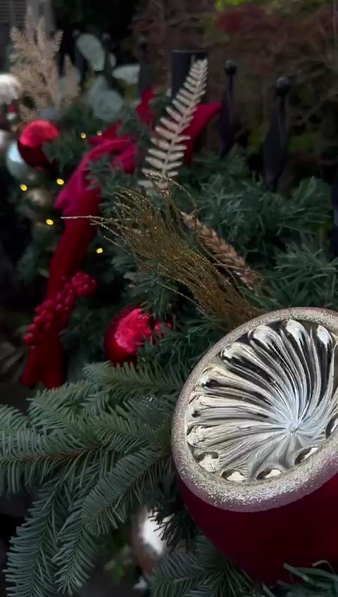 A close detail of red velvet ribbon, gilded fern and a large brushed silver
                    bauble set into a deep green garland.