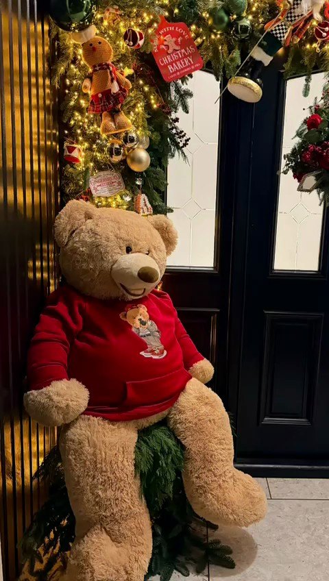 A dressed entrance with a giant seated teddy bear in a red hooded top beside a
                    black door framed by a lit garland with baubles and gingerbread figures.