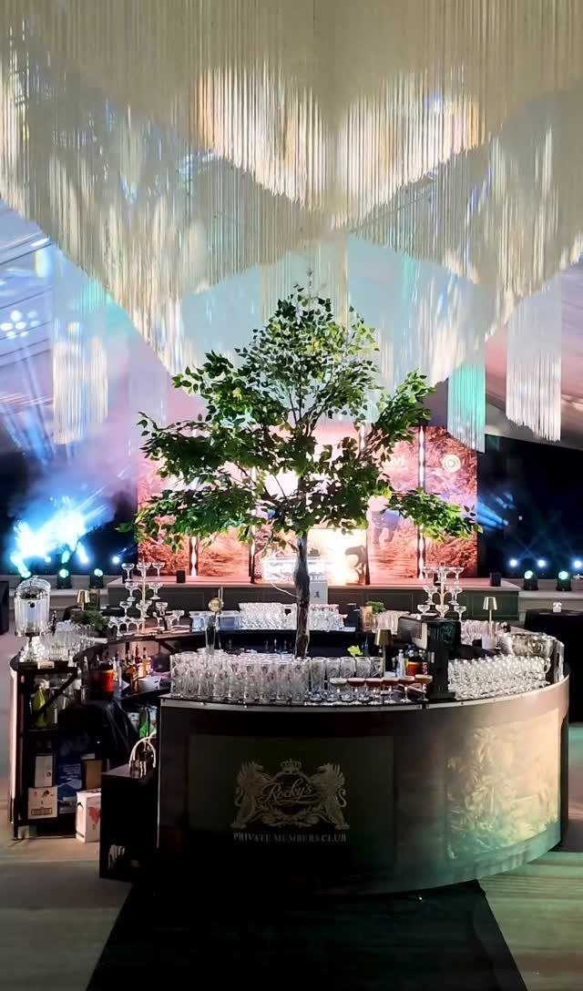 A large event space dressed for a party: a lit tree at the centre, a long
                    curved bar with glassware laid out, and a ceiling of hanging lights.